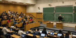 Starszy profesor w tweedowym garniturze piszący kredą na zielonej tablicy w nowoczesnej sali wykładowej, a studenci pracują na laptopach.